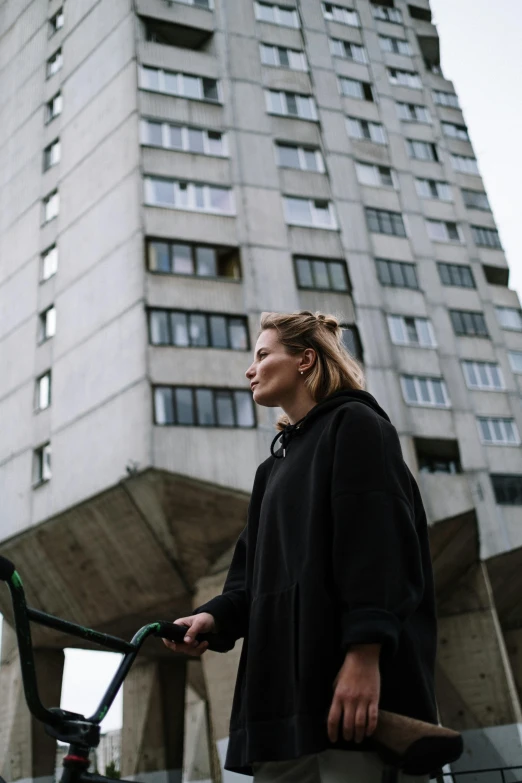 a woman standing in front of a tall building, by Emma Andijewska, wearing a black hoodie, mass housing, rollerblading, profile image