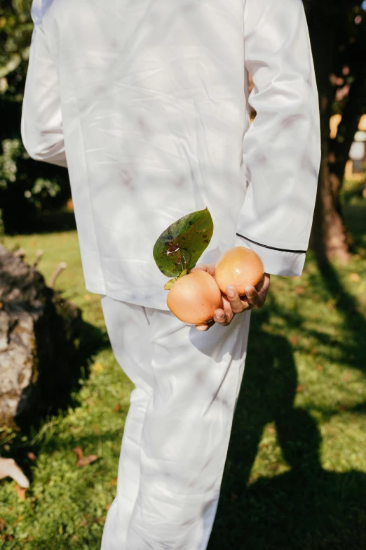 a man in a white suit holding an orange, by Julia Pishtar, unsplash, wearing white pajamas, leaves and simple cloth, pears, in style of lam manh