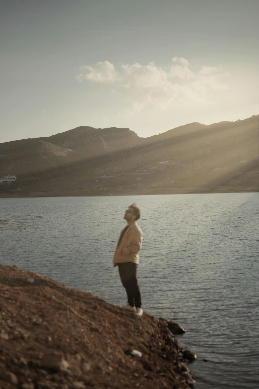 a person standing at the edge of a body of water, hills and ocean, surrounding cinematic light, pondering, doing a majestic pose