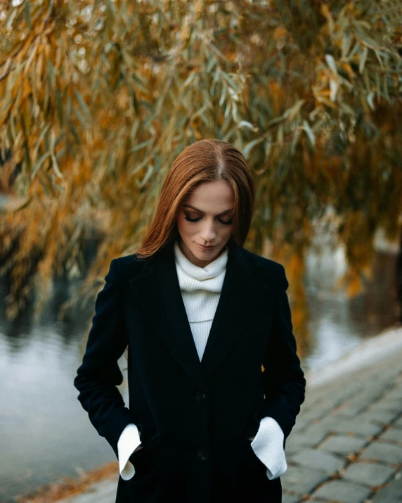 a woman in a black coat standing next to a body of water, wearing a suit and tie, during autumn, trending photo