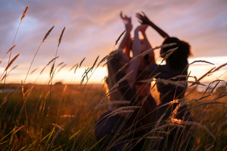 a couple of women standing next to each other in a field, by Jessie Algie, unsplash, land art, hands in air, standing in the grass at sunset, lachlan bailey, long grass in the foreground