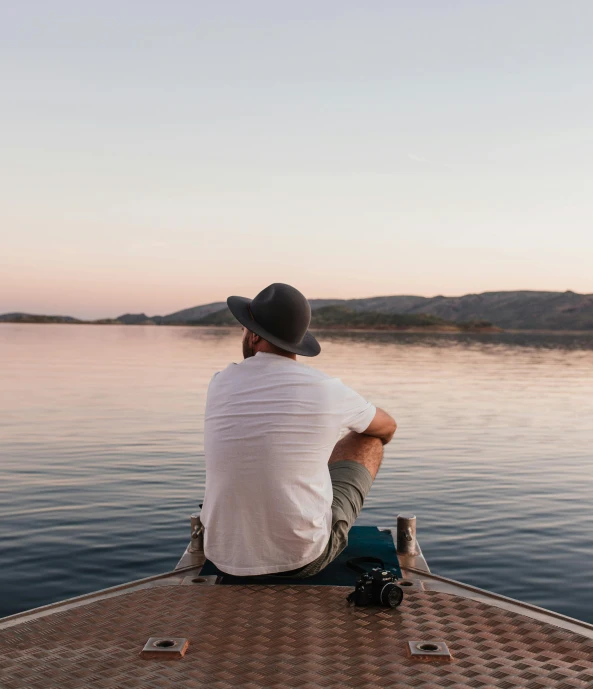 a man sitting on a boat looking out at the water, by Jessie Algie, pexels contest winner, wearing a travel hat, calm evening, in a vast serene landscape, a handsome