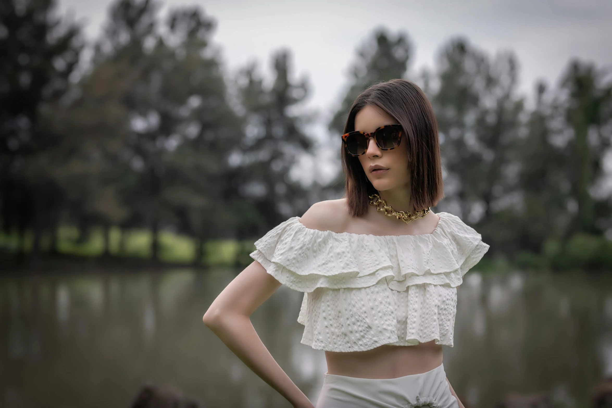 a woman standing in front of a body of water, an album cover, inspired by Elsa Bleda, pexels contest winner, renaissance, fashion model in sunglasses, at a park, ruffles, off the shoulder shirt