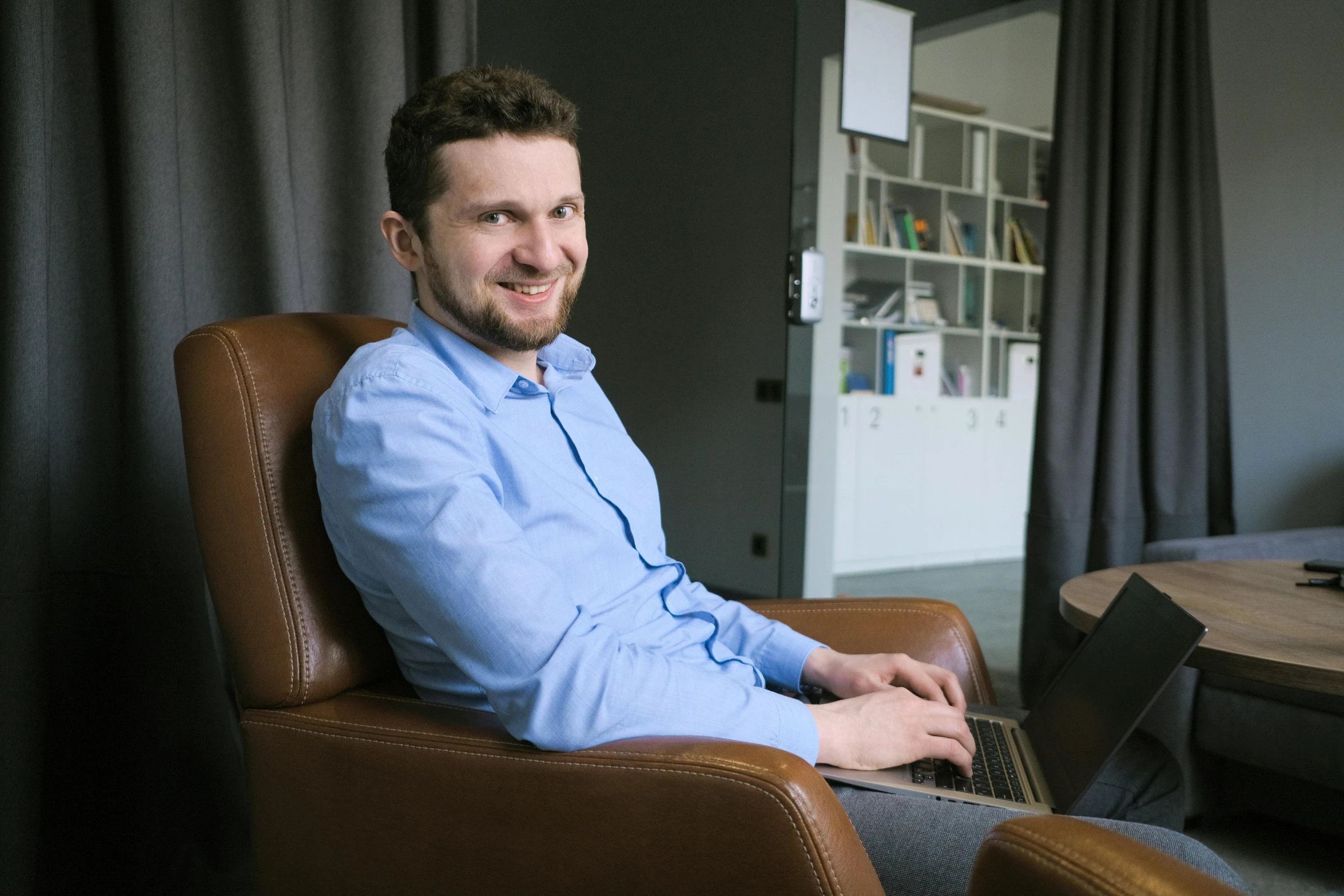 a man sitting in a chair with a laptop, by Adam Marczyński, smiling male, brown, in the office, slightly smirking