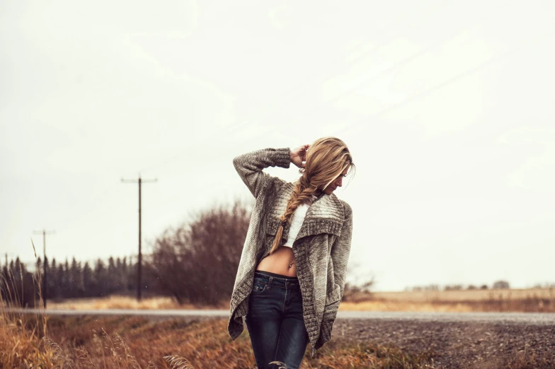 a woman standing in the middle of a road, trending on pexels, wearing a cardigan, loose braided hair, ( ( ( wearing jeans ) ) ), cold stormy wind