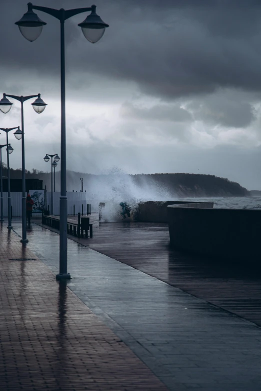 a person walking down a sidewalk next to a body of water, a picture, stormy seas, azores, lampposts, boardwalk