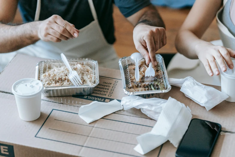 a couple of people sitting at a table with food, cardboard, rice, foil, hot food