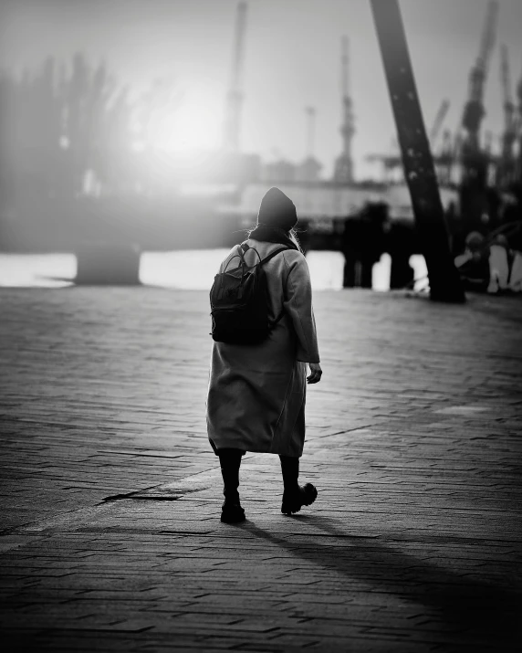 a black and white photo of a person with a backpack, by Tamas Galambos, unsplash contest winner, wearing a long coat, harbor, the girl and the sun, medium format. soft light