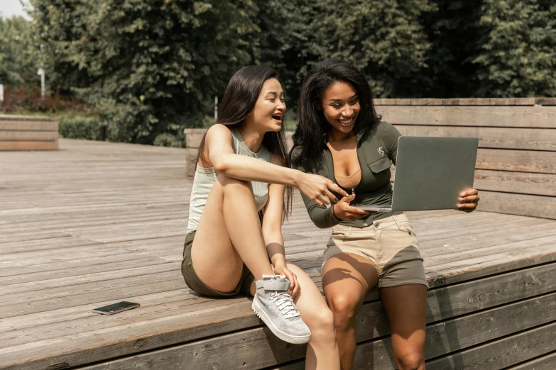 two women sitting on a bench looking at a laptop, trending on pexels, happening, tan skin a tee shirt and shorts, green and brown clothes, awarding winning, exposed thighs