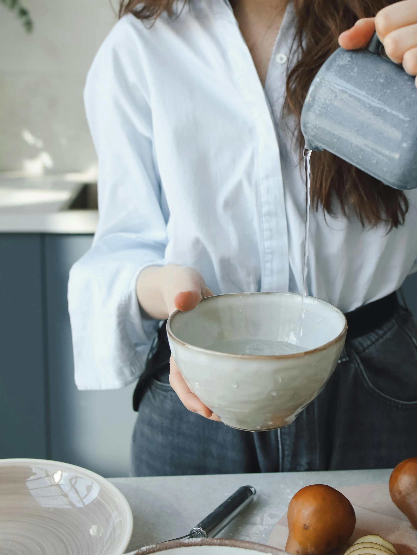 a woman is pouring something into a bowl, inspired by Kanō Naizen, trending on unsplash, soft grey and blue natural light, wearing a blouse, thumbnail, looking towards camera