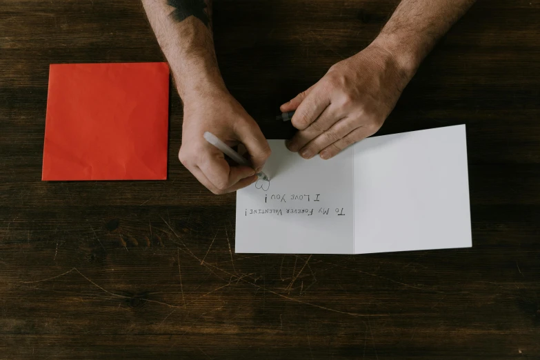 a man holding a pen and writing on a piece of paper, by Julia Pishtar, pexels contest winner, letterism, greeting card, red writing, cardboard, maintenance photo