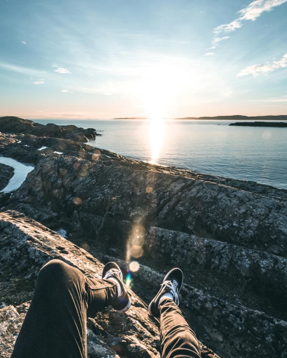 a person sitting on top of a rock next to the ocean, lying down, sun down, archipelago, legs taking your pov