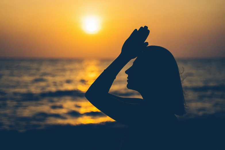 a woman standing on top of a beach next to the ocean, pexels contest winner, romanticism, praying at the sun, profile image, greeting hand on head, stylized silhouette