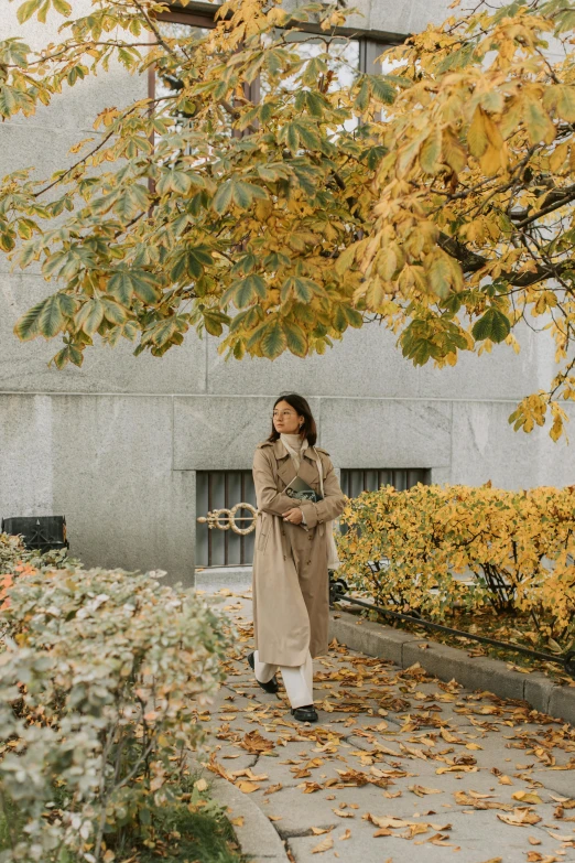 a woman standing under a tree in front of a building, trending on unsplash, visual art, wearing a long beige trench coat, autumn season, vancouver, low quality photo