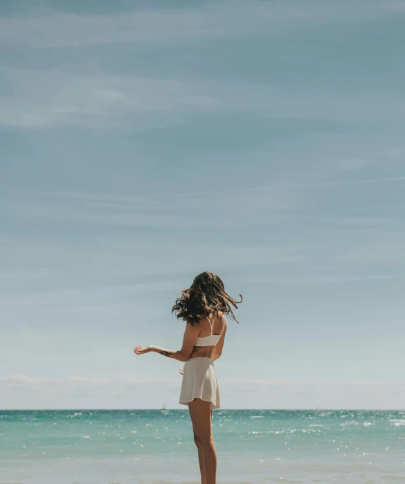 a woman standing on a beach next to the ocean, unsplash contest winner, minimalism, wearing a camisole and shorts, swirling around, clear blue skies, looking from behind