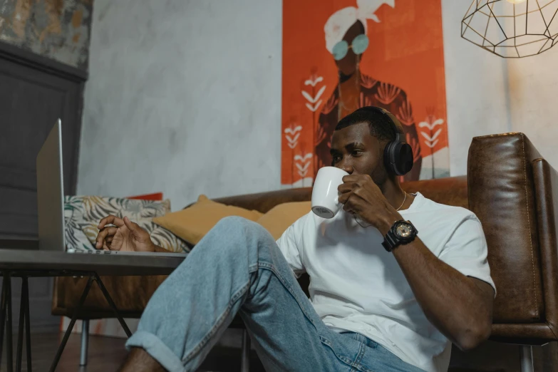 a man sitting on a couch with headphones on, by Adam Marczyński, trending on pexels, afrofuturism, is ((drinking a cup of tea)), two buddies sitting in a room, low quality photo, lounge background