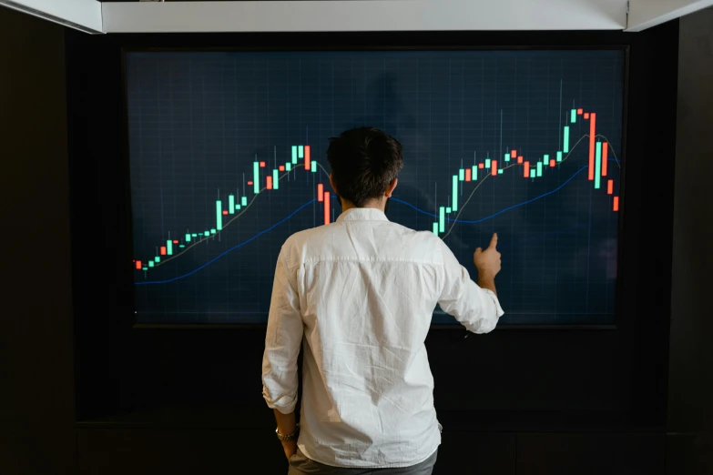 a man standing in front of a screen with a stock chart on it, trending on unsplash, 🦩🪐🐞👩🏻🦳, teaching, 15081959 21121991 01012000 4k, lachlan bailey