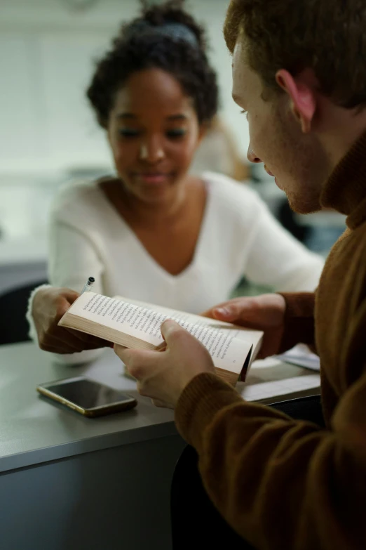 a couple of people that are sitting at a table, holding a book, varying ethnicities, in a classroom, light glow