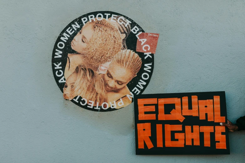 a person standing next to a wall with stickers on it, trending on pexels, black arts movement, woman holding another woman, with a ponytail, protest movement, white and orange breastplate