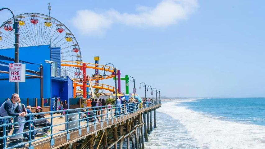 a group of people standing on top of a pier next to the ocean, ferris wheel, it's californication, brightly colored buildings, profile image