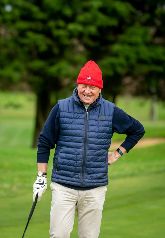 a man standing on top of a green field holding a golf club, wearing a red gilet, beanie, ian callum, profile image