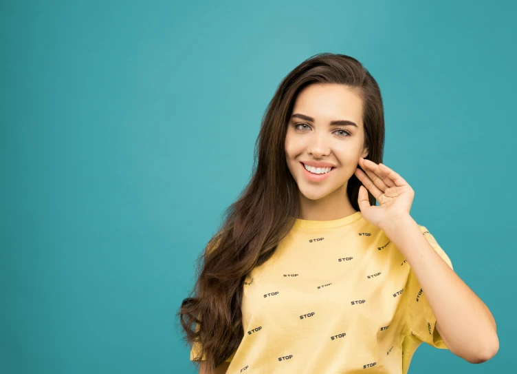 a woman holding a cell phone up to her ear, trending on pexels, antipodeans, wearing a modern yellow tshirt, cyan photographic backdrop, straight teeth, isabela moner