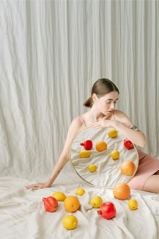 a woman sitting in front of a mirror surrounded by fruit, soft round features, yellow and red, in front of white back drop, soft aesthetic