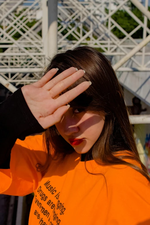 a woman in an orange shirt covering her eyes, an album cover, inspired by Elsa Bleda, trending on pexels, realism, trending on r/streetwear, hand gesture, 🚿🗝📝, on a bridge