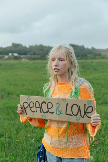 a woman holding a sign that says peace and love, an album cover, unsplash, blonde swedish woman, young teen, movie still frame, camp