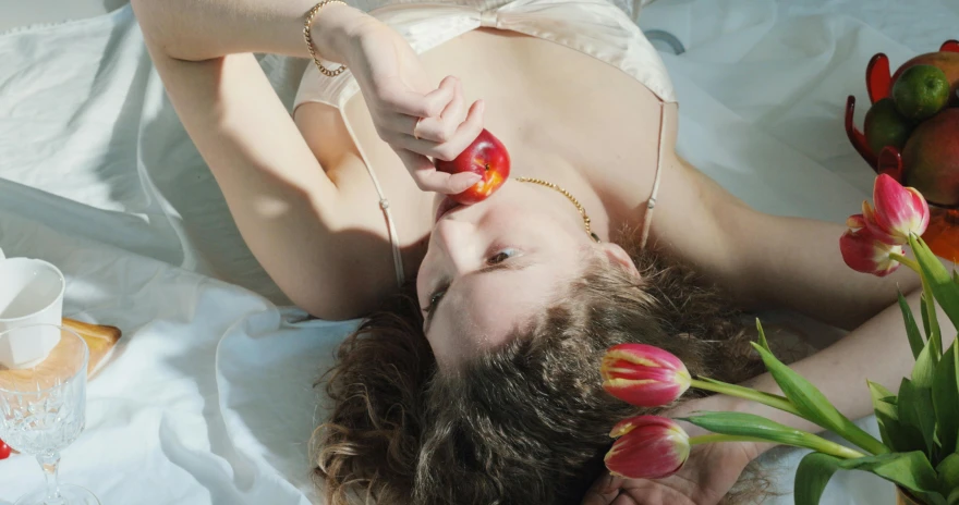 a woman laying on top of a bed next to flowers, inspired by Elsa Bleda, pexels contest winner, magic realism, she is eating a peach, red apple, hans bellmer and nadav kander, holding an epée