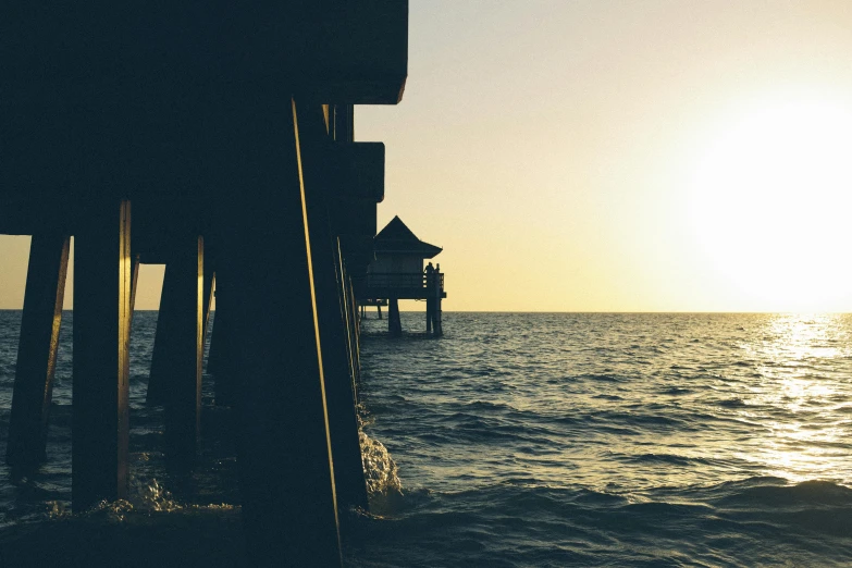 the sun is setting over the water near a pier, by Niko Henrichon, pexels contest winner, houses on stilts, vintage photo, ferrofluid oceans, view from side