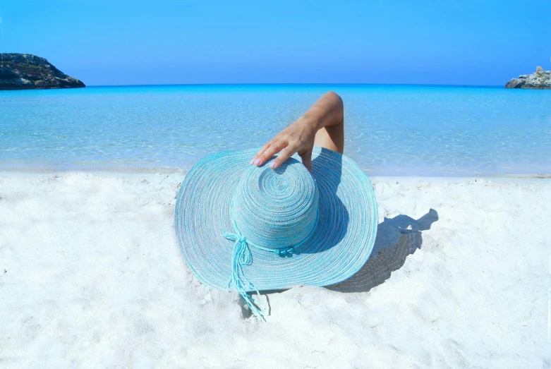 a woman sitting on top of a sandy beach next to the ocean, blue fedora, blue background colour, light blue skin, hand holding cap brim