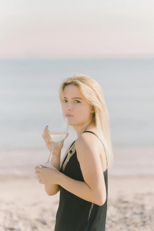 a woman standing on a beach holding a glass of wine, a girl with blonde hair, profile image, vine, portrait image