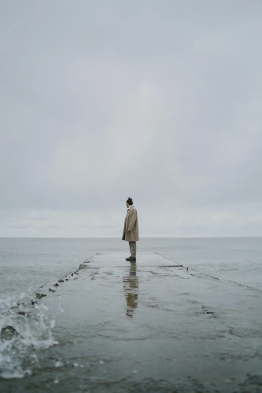 a man standing in the middle of a body of water, unsplash, minimalism, woman is in a trenchcoat, near a jetty, overcast gray skies, ignant