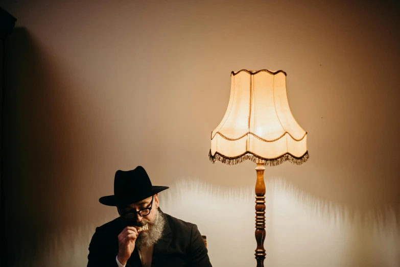 a man sitting at a table in front of a lamp, by Will Ellis, unsplash, magical realism, wearing a fedora, orthodox, sholim, floor lamps