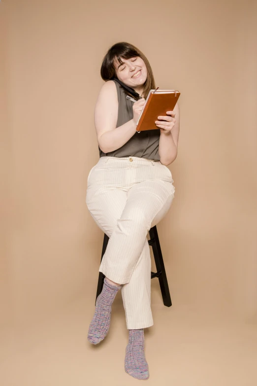 a woman sitting on a stool reading a book, by Carey Morris, nonbinary model, slightly overweight, holding a clipboard, brown pants