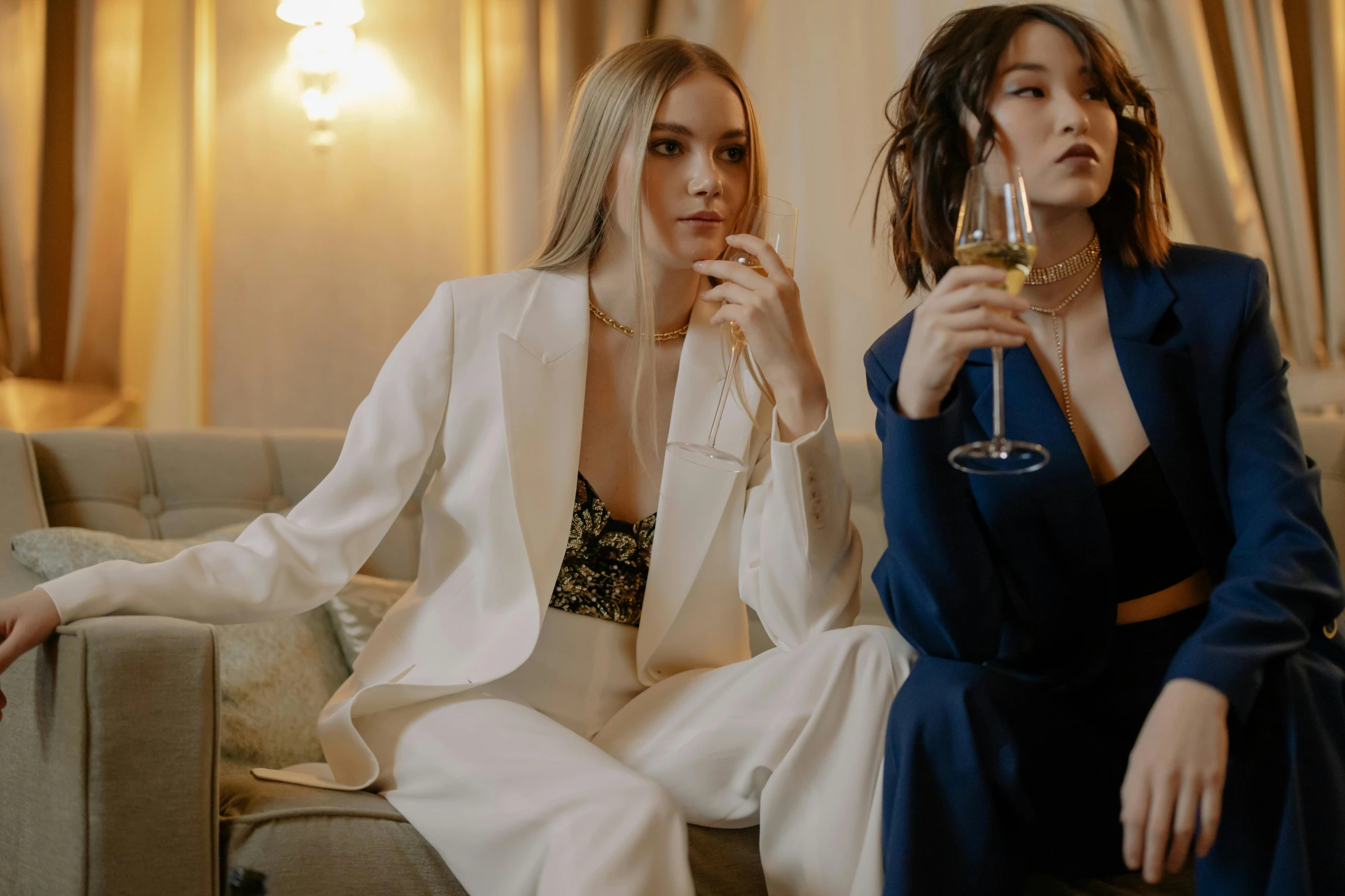 two women sitting on a couch holding wine glasses, by Emma Andijewska, trending on pexels, art nouveau, three piece suit, white russian clothes, party, marble and hint gold