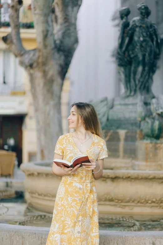 a woman standing in front of a fountain holding a book, happening, wearing yellow floral blouse, exploration, in town, robe