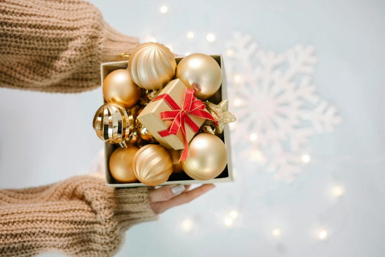a person holding a box of christmas ornaments, by Elaine Hamilton, pexels contest winner, beige and gold tones, avatar image, ornament, square