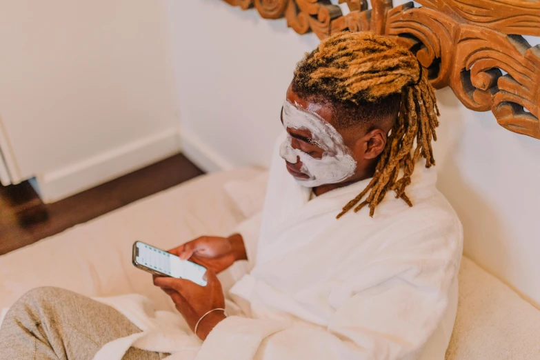 a person sitting on a bed using a cell phone, trending on pexels, afrofuturism, white face paint, spa, a man wearing golden mask, with textured hair and skin