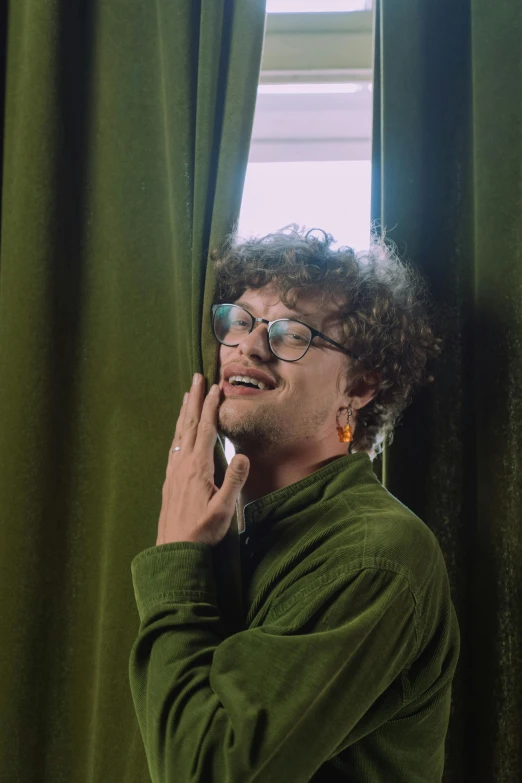 a man standing in front of a window talking on a cell phone, an album cover, by Jacob Toorenvliet, trending on pexels, renaissance, wearing small round glasses, he has short curly brown hair, wearing a green sweater, holding a pudica pose