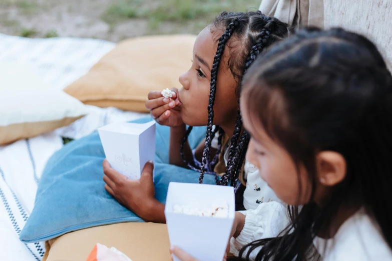 two little girls sitting on a blanket eating popcorn, by Emma Andijewska, pexels contest winner, people outside eating meals, cardboard, close up to the screen, avatar image