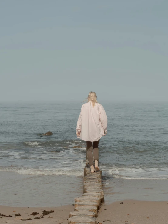 a woman standing on top of a beach next to the ocean, an album cover, unsplash, happening, pastel pink robes, girl with short white hair, lonely human walking, low quality footage