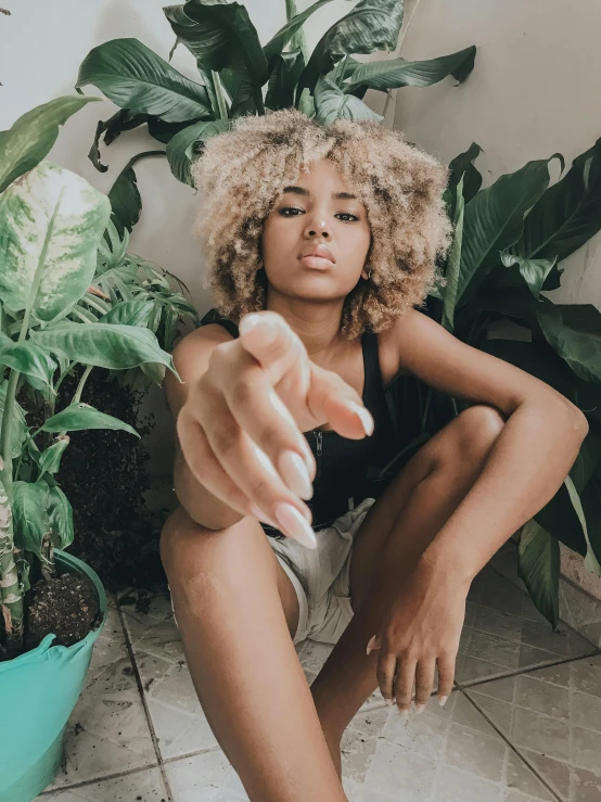 a woman sitting on the floor with a remote control in her hand, inspired by Elsa Bleda, trending on unsplash, afrofuturism, woman made of plants, short blonde afro, giving the middle finger, lush surroundings