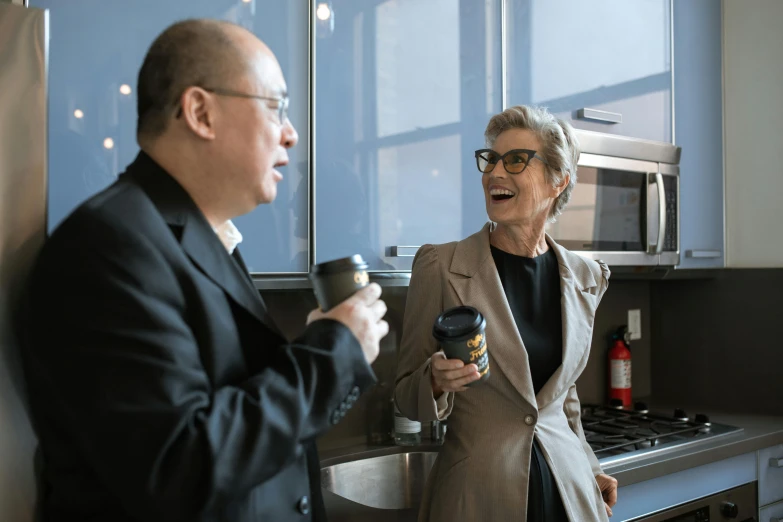 a man and a woman standing in a kitchen, in suit with black glasses, drinking a cup of coffee, old lady screaming and laughing, jen atkin