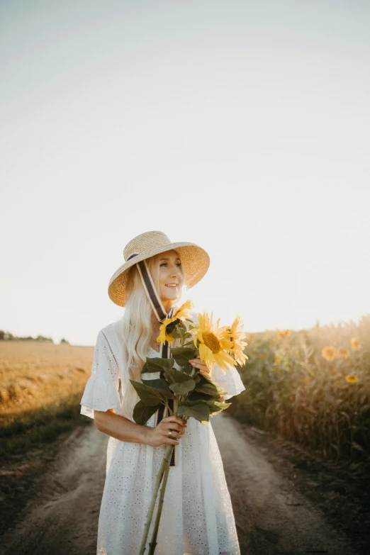 a woman standing on a dirt road holding a bunch of sunflowers, inspired by Sun Long, pexels contest winner, minimalism, wearing a cute hat, delightful surroundings, a beautiful woman in white, blonde woman