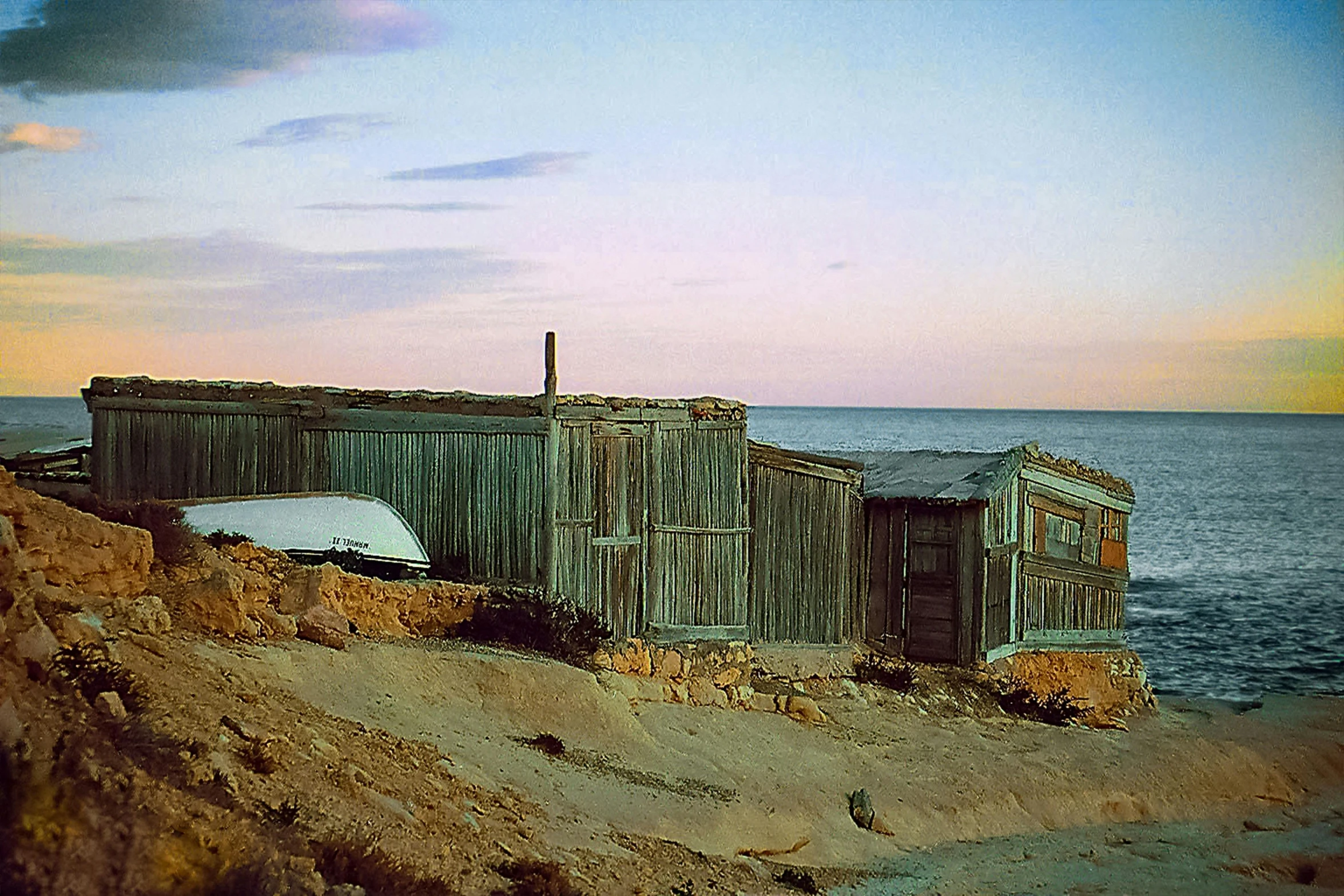 a boat sitting on top of a sandy beach, a colorized photo, by Morris Kestelman, unsplash, australian tonalism, rustic stone cabin in horizon, wooden houses, photo taken on fujifilm superia, mining outpost