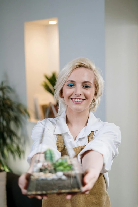 a woman holding a tray with a plant in it, a character portrait, by Adam Marczyński, pexels contest winner, bartending, evanna lynch, head and shoulders shot, white apron