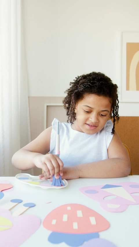 a little girl that is sitting at a table, holding a paintbrush, soft shapes, keter class, diecut