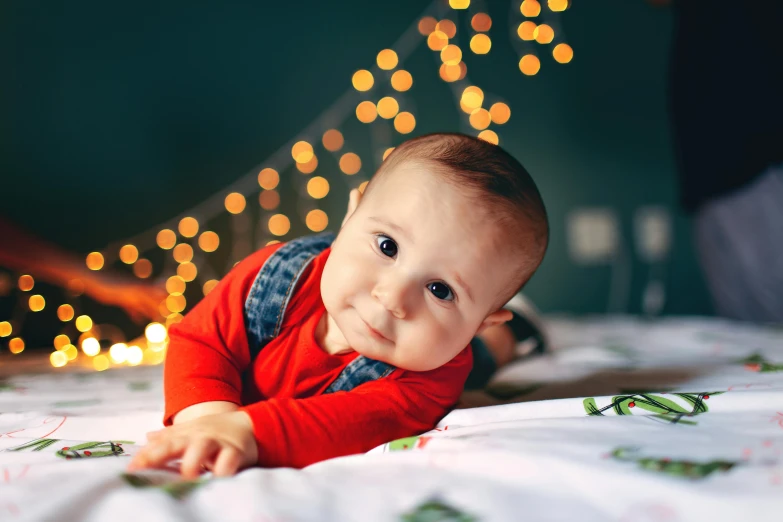 a baby laying on top of a bed next to a christmas tree, pexels contest winner, light stubble with red shirt, string lights, looking towards camera, gif